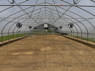The image is the inside of an empty high tunnel greenhouse.  The grey, partially transparent walls come to a teardrop peak and are rounded down to the ground.  There is an orange dirt floor.