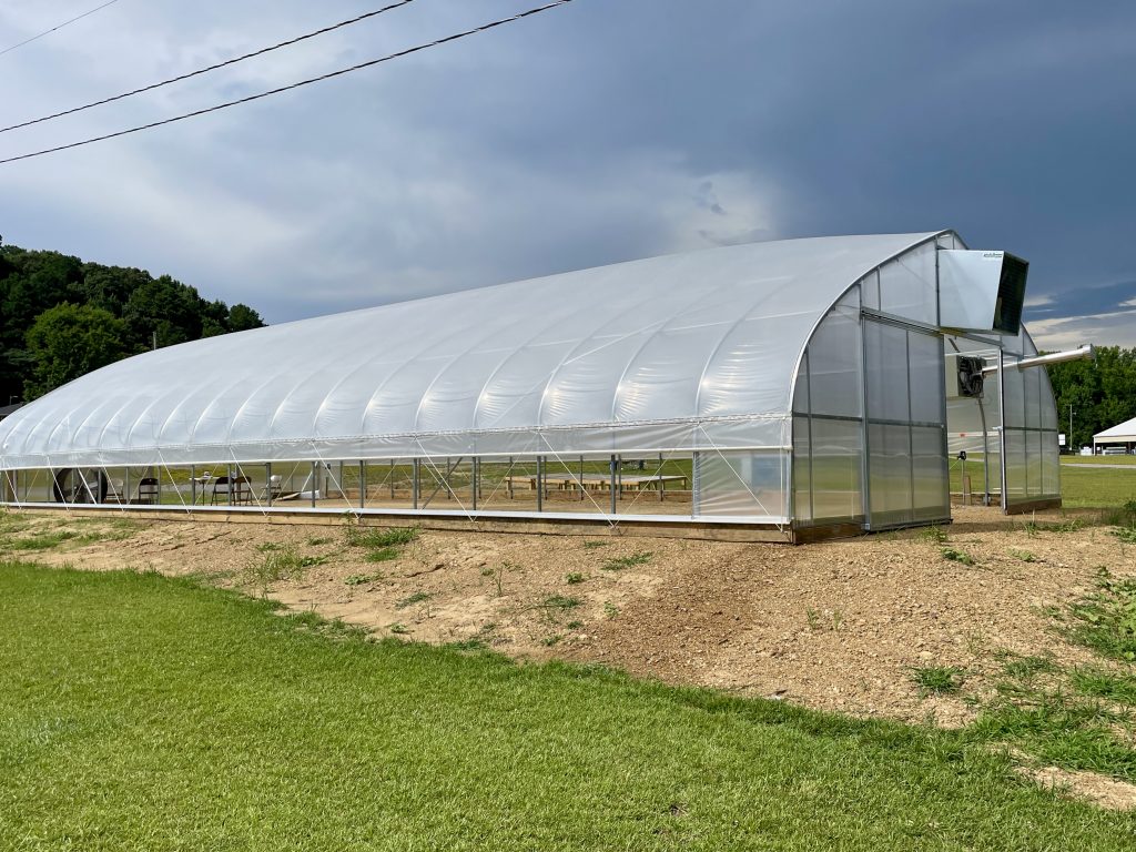 A new high tunnel greenhouse with grey film stretched across the roof supports with fans and vents and large doors on both ends.