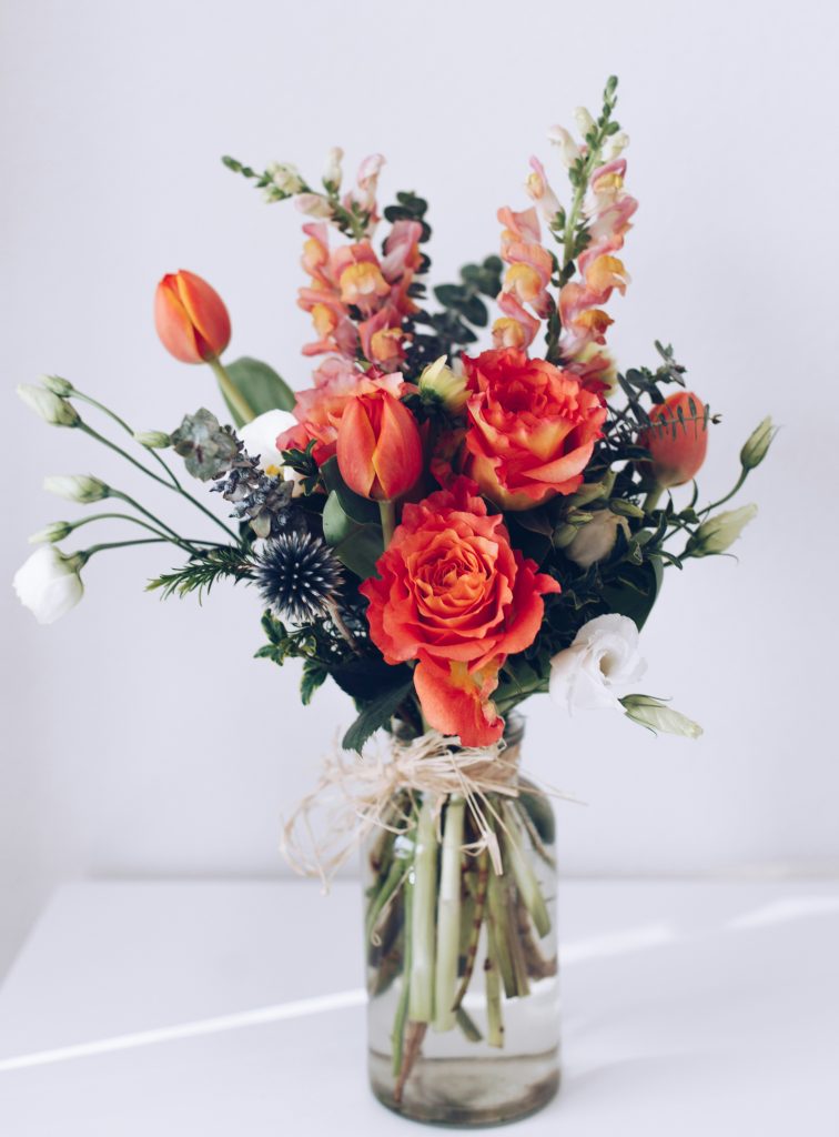 A clear mason jar of dark coral-colored roses with lighter coral-colored snapdragons and tulips. Sprigs of Eucalyptus are positioned in the back and on the sides and with white daisies emerge from both sides. A thin raffia bow is tied around the neck of the jar.